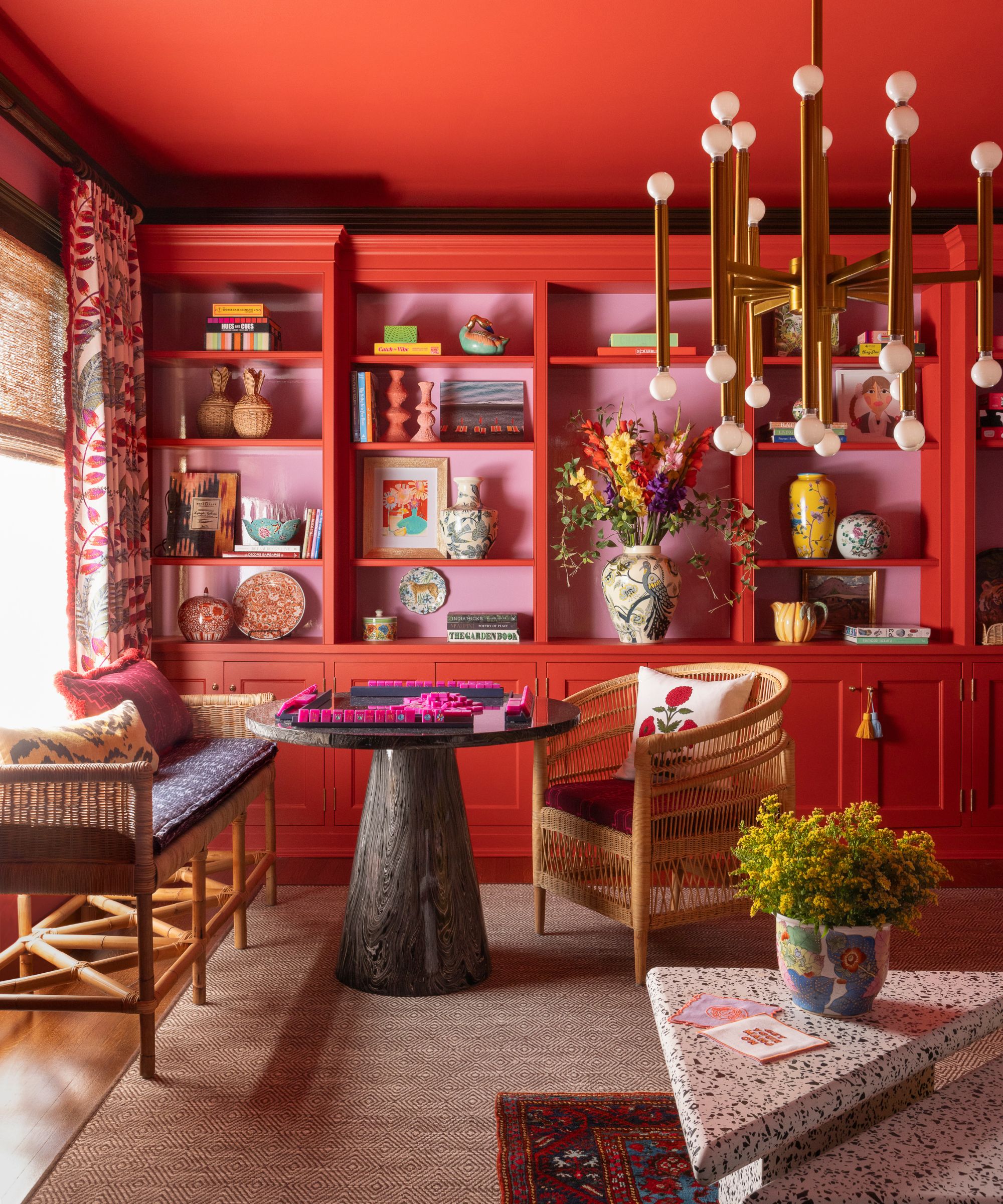 Living room with bright red painted ceiling and an open bookcase painted pink and red, a jute rug, two rattan chairs, a marble round table, and a terrazzo coffee table