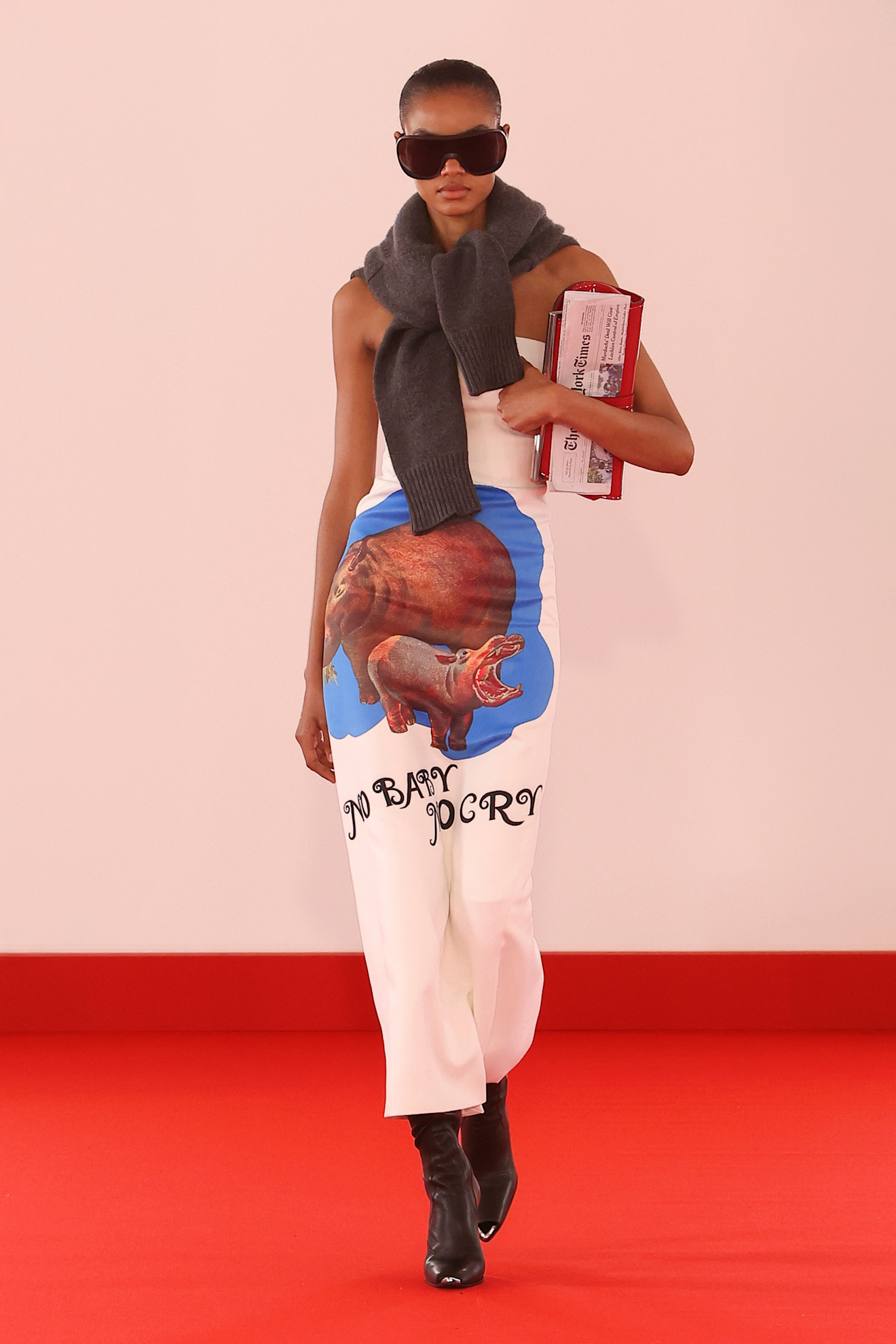 Layering staples for spring 2026 is shown in a photo from Brandon Maxwell's runway show, with a model walking while wearing a gray sweater tied around the shoulders, worn with a white tube top, a white printed pencil skirt, black knee-high boots, oversized aviator sunglasses, and a newspaper-shaped clutch bag