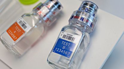 Two clear bottles with silver caps are lying flat against a white surface. One bottle has an orange label with the word test on it and the other has a similar label but is blue.