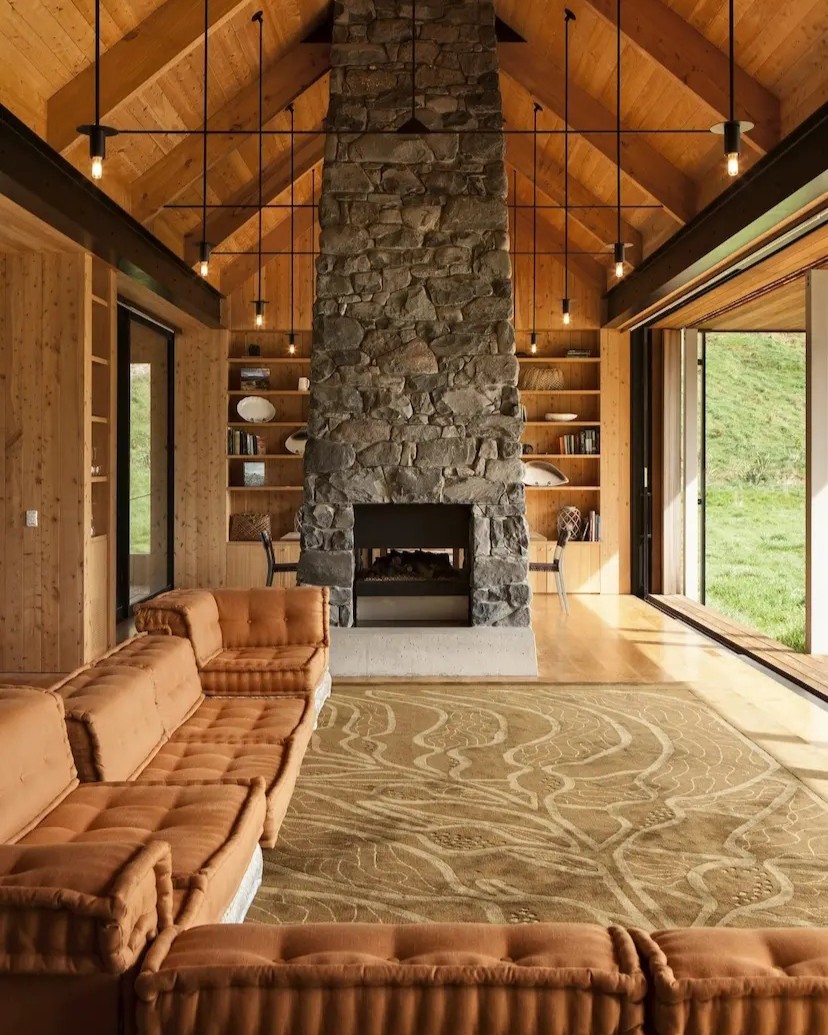 A 1970s-inspired mountain lodge is filled with a earthy modular couch, sandy rugs, wooden floors, and a modernist fireplace carved from rock and towering in a naturally lit room.