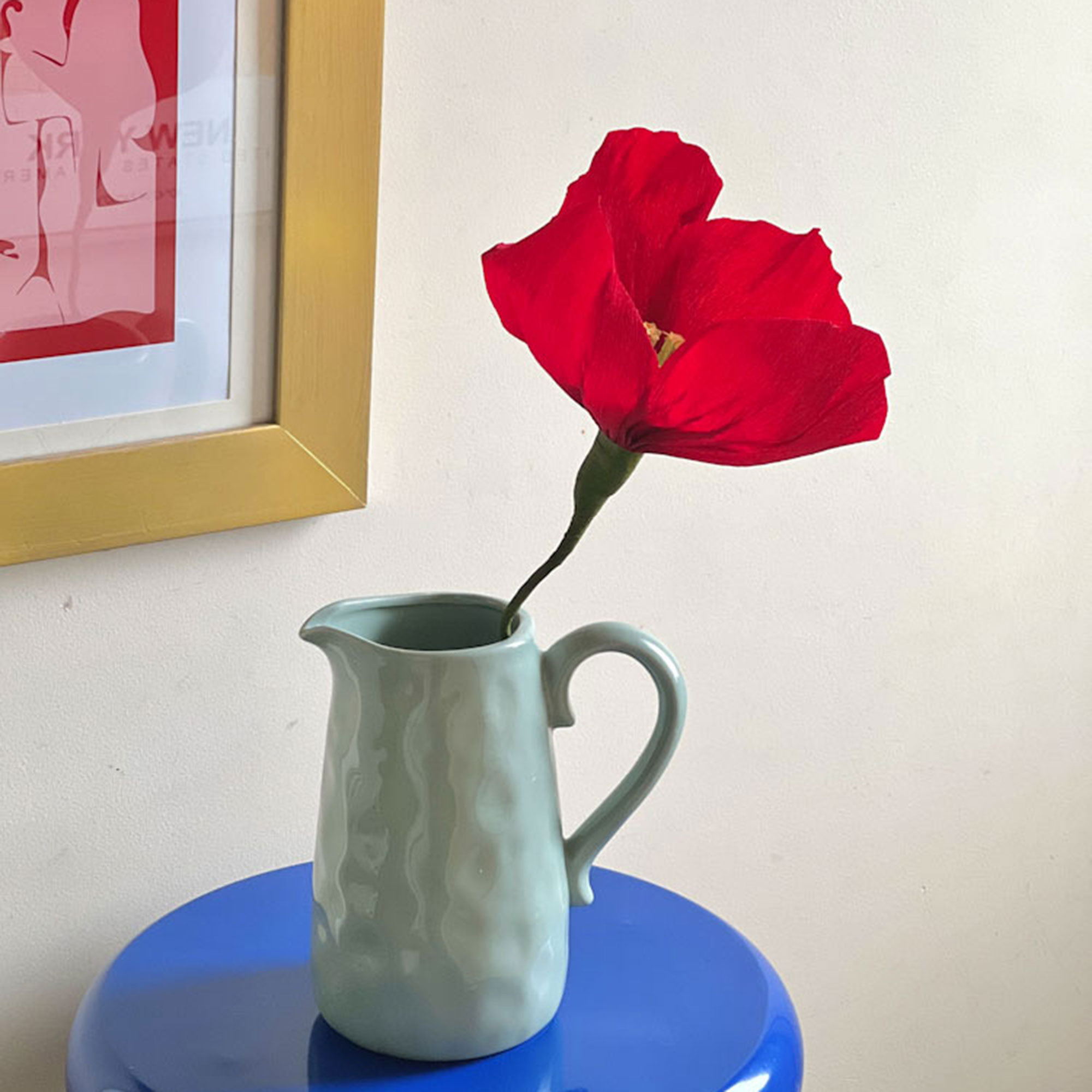 Red flower in green jug on bright blue side table
