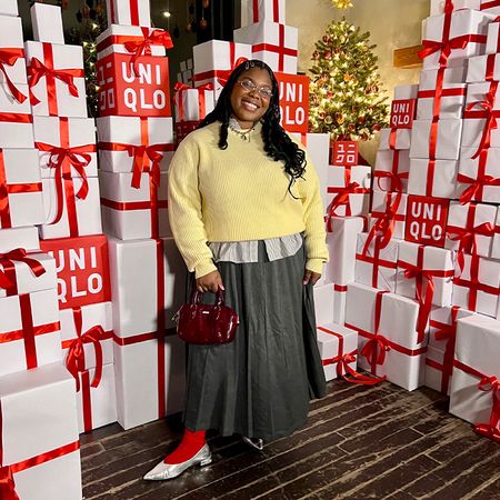 chichi wears yellow sweater layered over button down striped shirt, a long pleated gray skirt, red socks, silver pointed flat shoes, and a deep red brahmin handbag