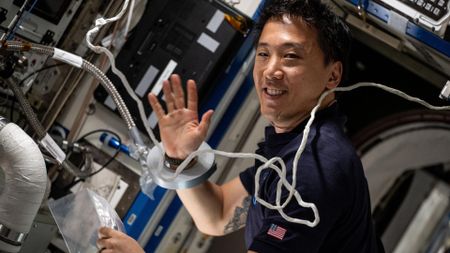 a man smiles and waves to a camera in a crowded laboratory full of wires and computer monitors