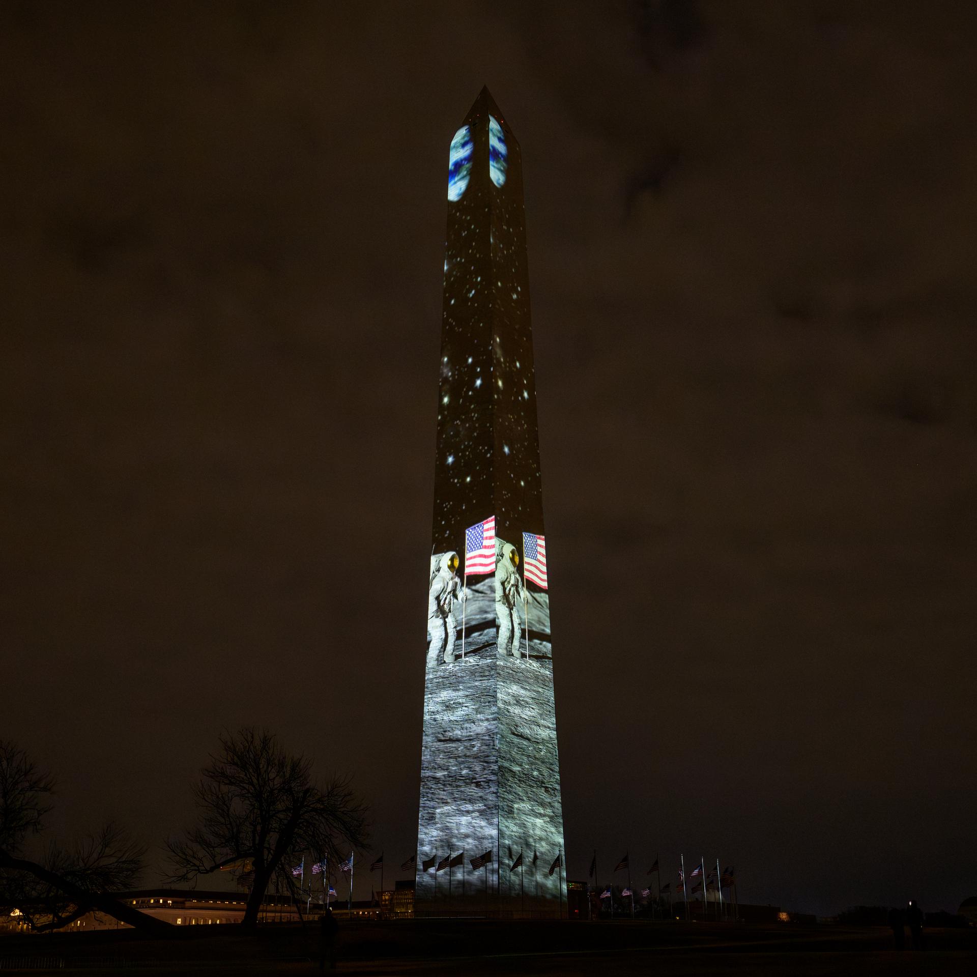 A tall obelisk-shaped monument is illuminated with projectors to produce scenes of rockets, astronauts on the moon, and the numbers 250 vertically down the building