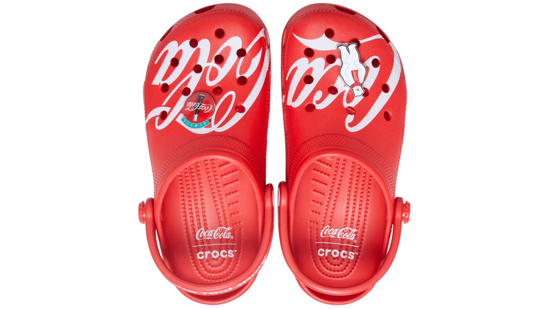 A top-down view of a pair of bright red Crocs featuring the white Coca-Cola script logo printed across the toe box, complete with a round Coca-Cola bottle Jibbitz charm on one shoe and a polar bear charm on the other.
