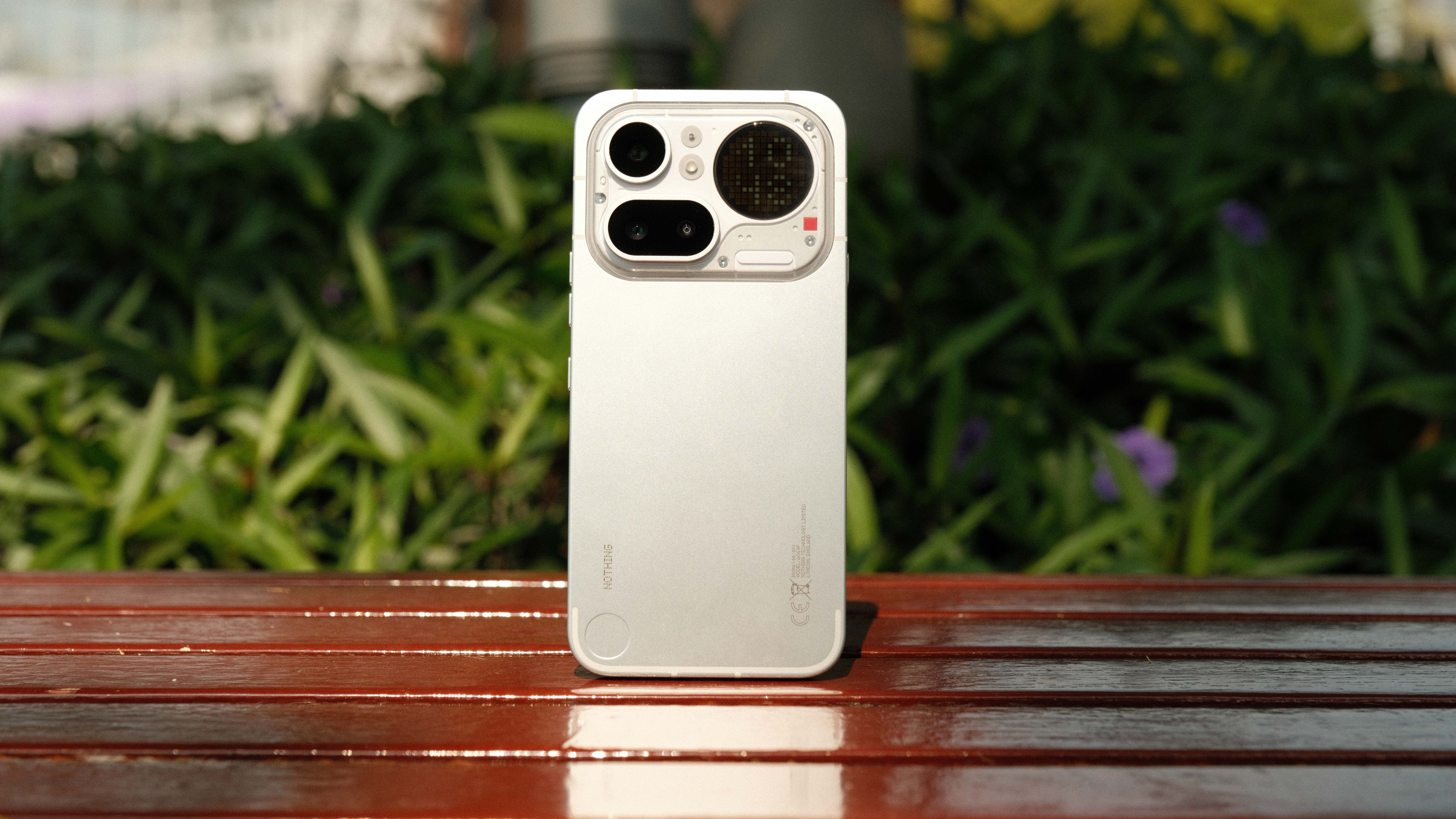 A vertical shot of the back of a light gray smartphone standing upright on a wooden surface. The design features a large, transparent rectangular camera section at the top and minimalist branding at the bottom, set against a soft-focus background of green garden foliage.