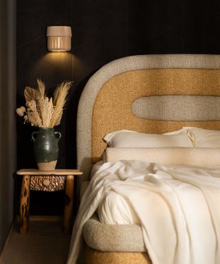 A dark bedroom with a brown and beige textured bedframe, a side table with a green vase, and a small sconce