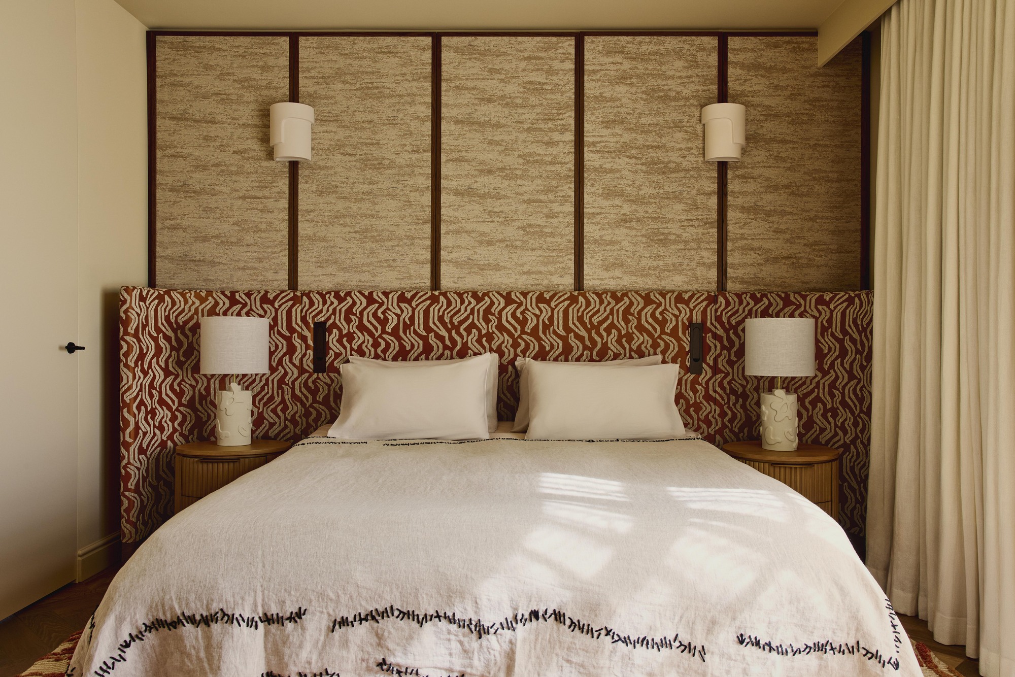 Image of a red-and-white patterned extended headboard in a beige room where one wall is upholstered. The bed has white bedding with black stitching around the bottom.
