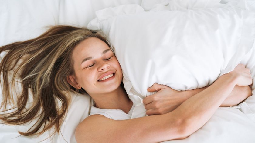 A woman lays in bed, smiling with her eyes closed, while holding her pillow.