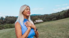 A woman stands in a field, with her hands held against her heart, eyes closed, and a gentle smile on her face.