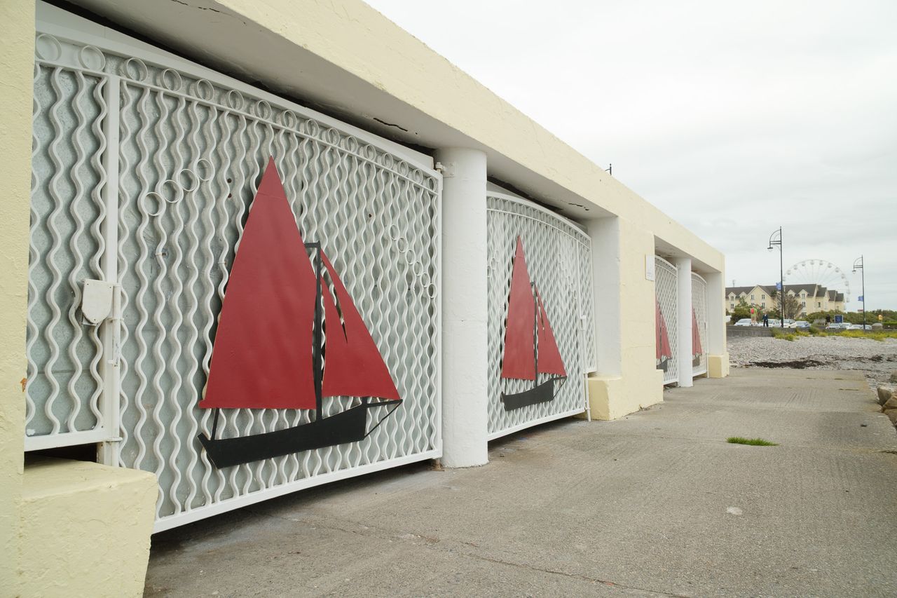 Red sailboat decorations mounted on white metal gates along a seaside promenade, with a Ferris wheel in the distance.