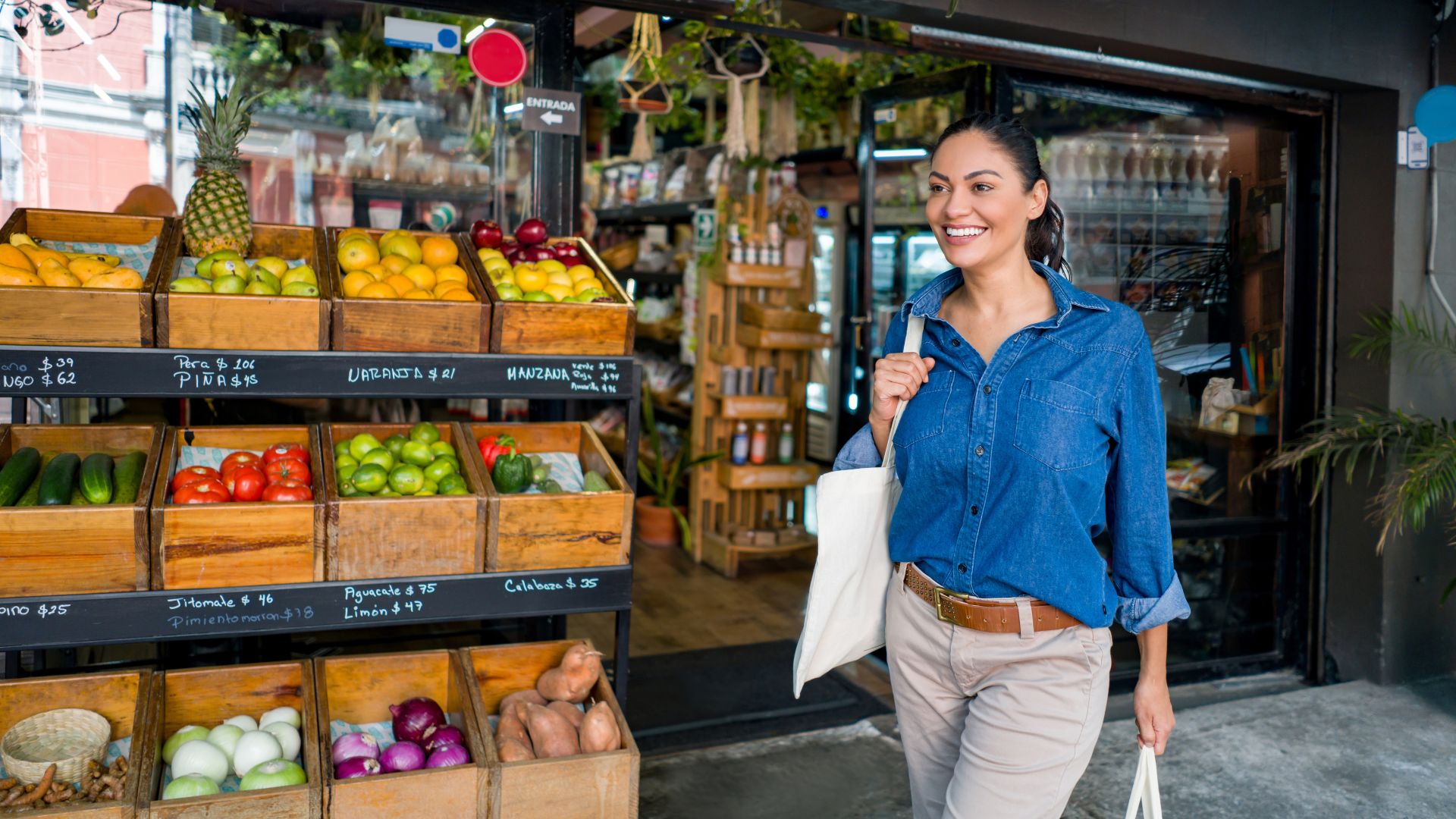A happy young woman with a white tote bag walks out of a grocery store in the morning.