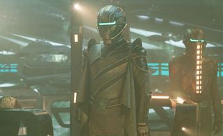 a humanoid alien wearing a brozne spacesuit-like costume stands on the bridge of a spaceship