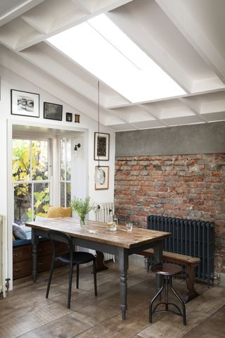 Dining area with wooden table, black metal radiator, wooden floor, red brick wall and sloped white ceiling with skylight