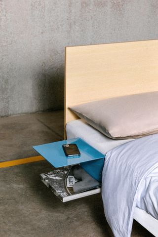 Detail of the bed showing the ash headboard and side table in blue aluminium. The bed is seen with beige pillow and light blue covers, and a phone is resting on the bedside table