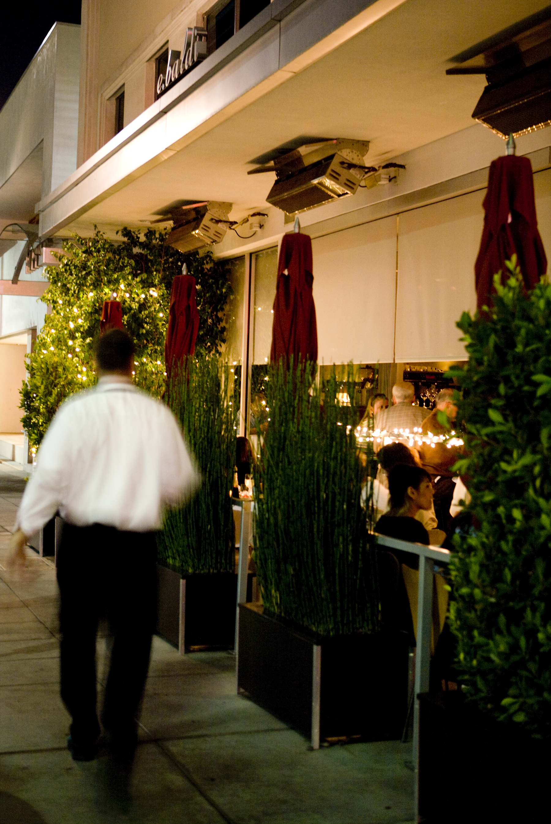 A restaurant's exterior features lush planters illuminated by trails of fairylights, people dining outside, and waiters on the move.