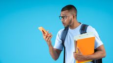 Young man look at this phone in surprise, while holding his notebooks and carrying a blue backpack on his back, on a blue background