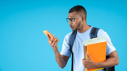 Young man look at this phone in surprise, while holding his notebooks and carrying a blue backpack on his back, on a blue background