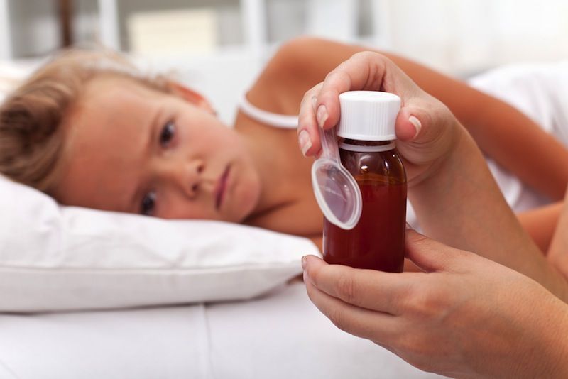 A little girl lays in med while her mom prepares a dose of medicine for her.