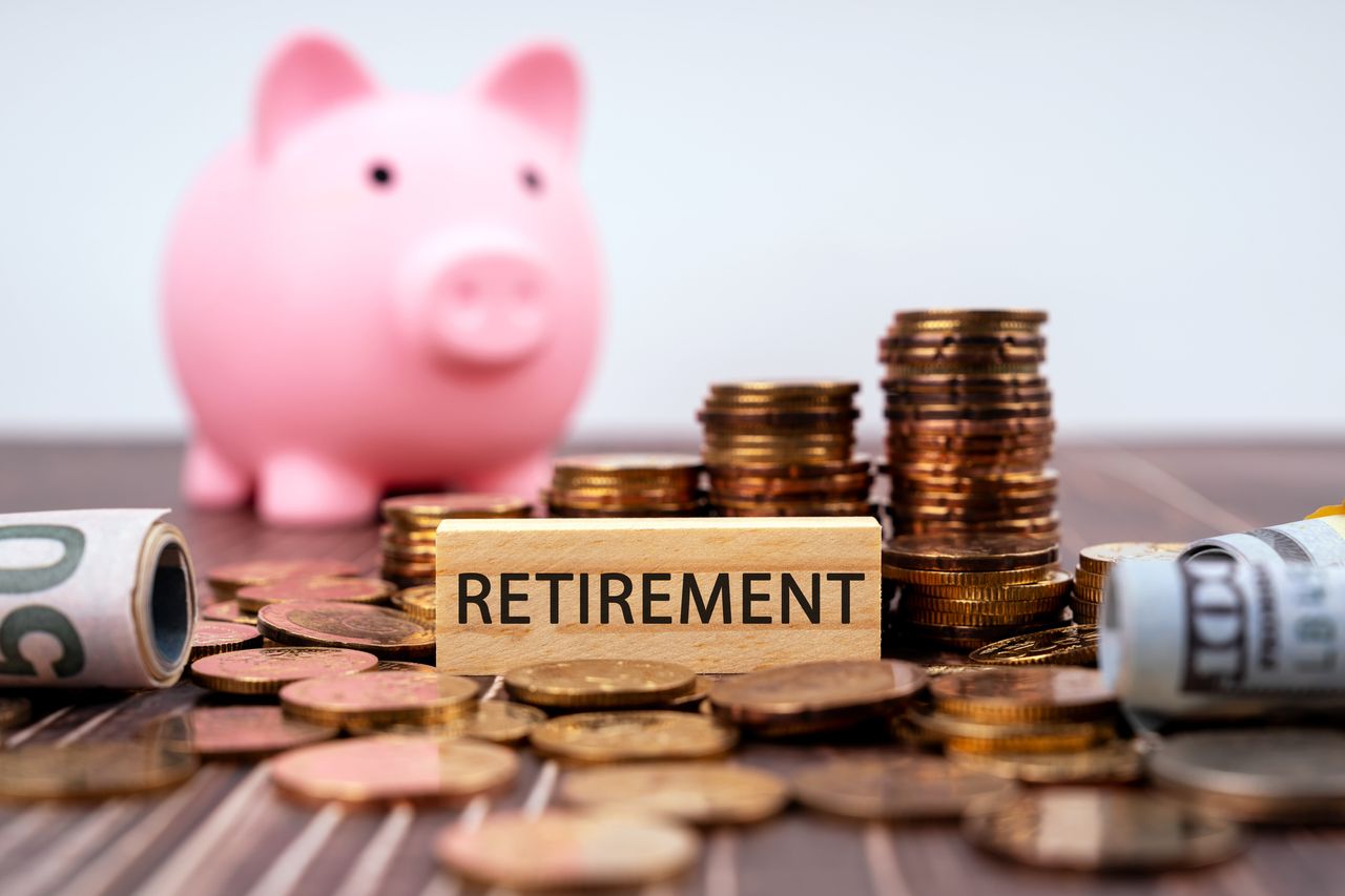 Wooden block with the word &quot;retirement&quot; sits on table with cash, coins and a piggybank.