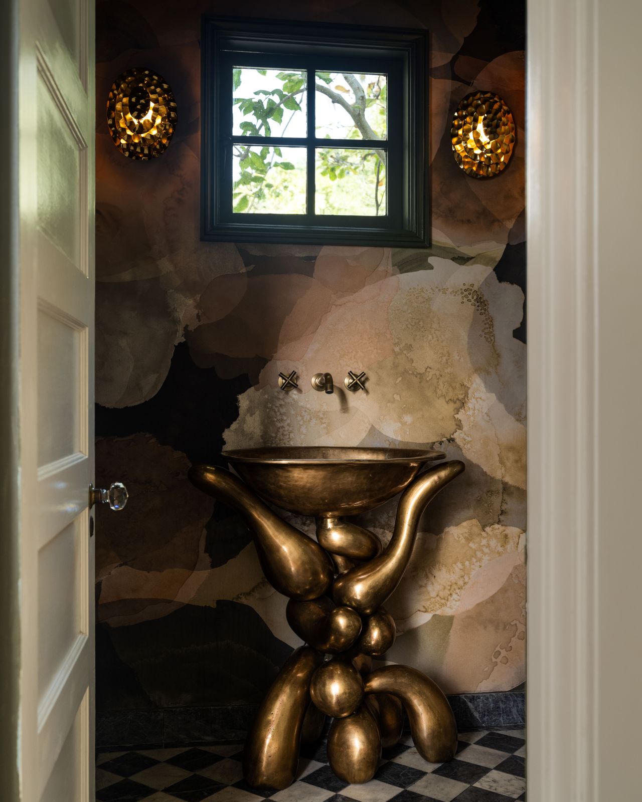 A lavishly decorated bathroom features a golden, sculptural sink and matching lights, tiled floors, and mural-like walls.
