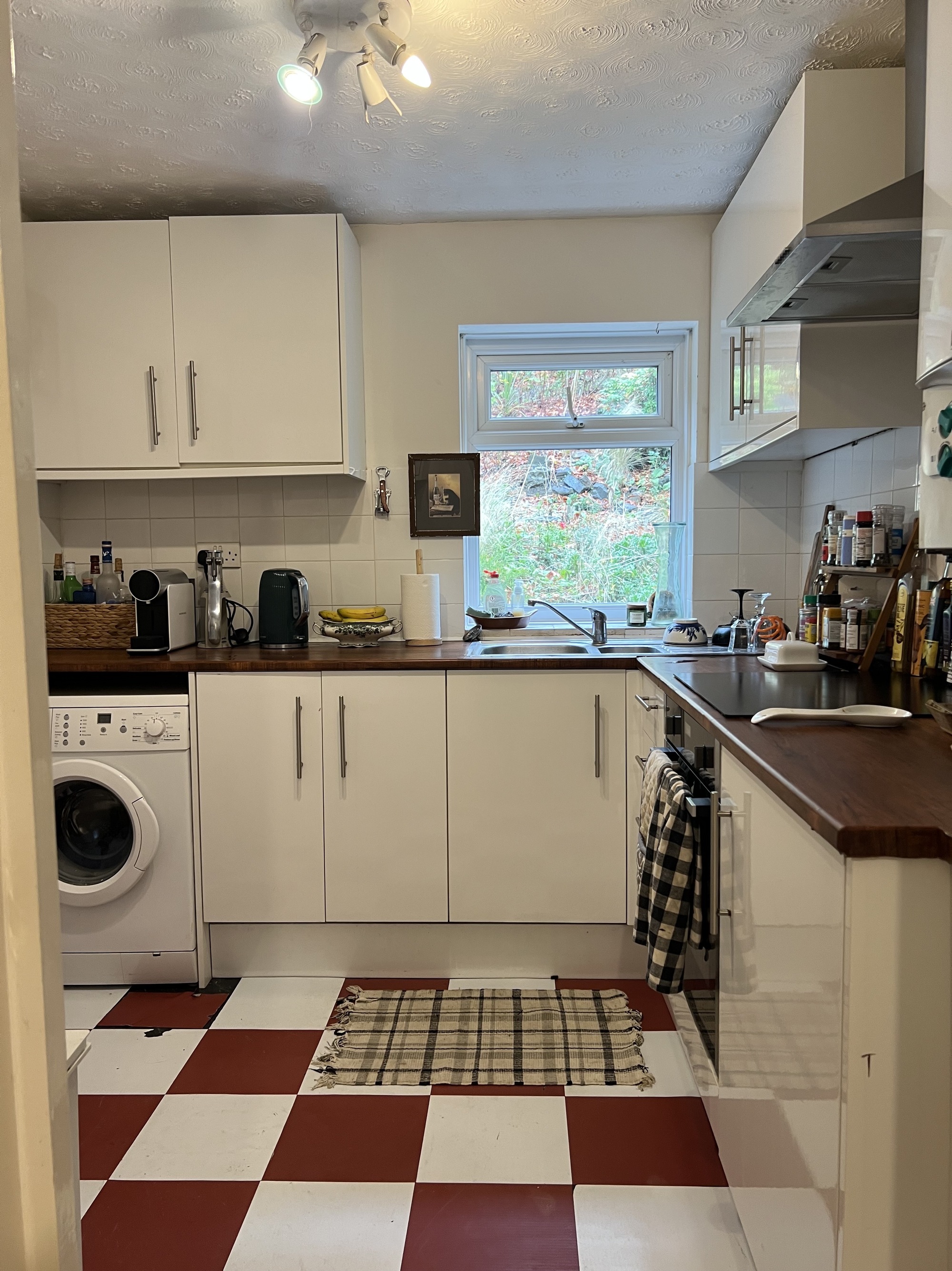 Image of a small apartment kitchen with red and white floor tiles and a faux wooden countertop. There is white cabinetry and a washing machine unit in the space.