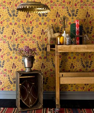 An entryway with yellow floral wallpaper, a wooden console table with books on top of it, and a dark wooden table with a vase of flowers on top of it