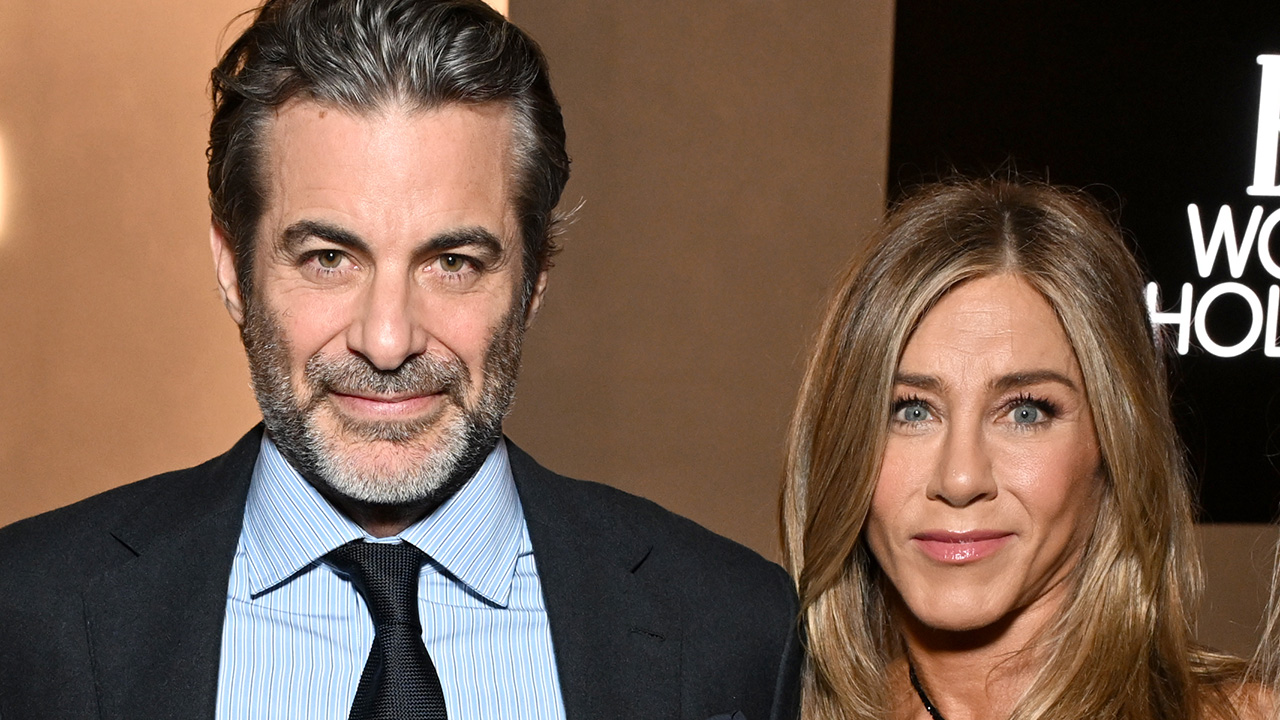Jennifer Aniston and Jim Curtis dressed up in a dress and suit and tie and staring straight at the camera.