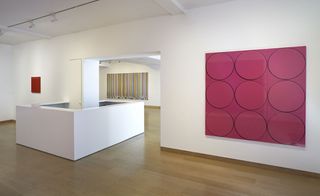 A gallery with a painting with a pink background and nine black circles on it on the wall.
