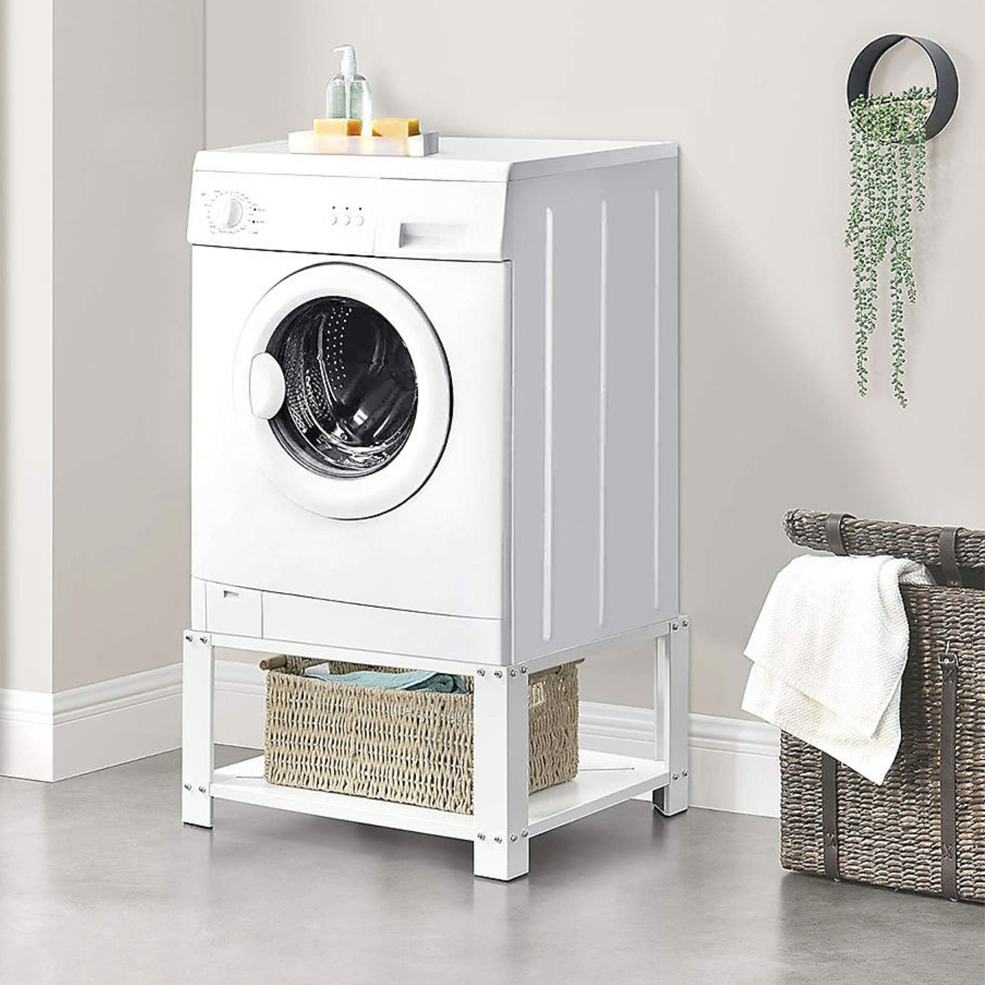 A white front-loading washing machine is elevated on a metal stand with a wicker basket stored below.
