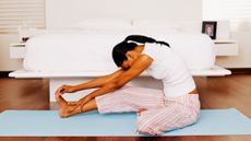 A woman performs yoga in a bedroom. She is on a yoga mat at the foot of her bed, one leg stretched out in front and the other bent on the floor, while she reaches forward with her torso.