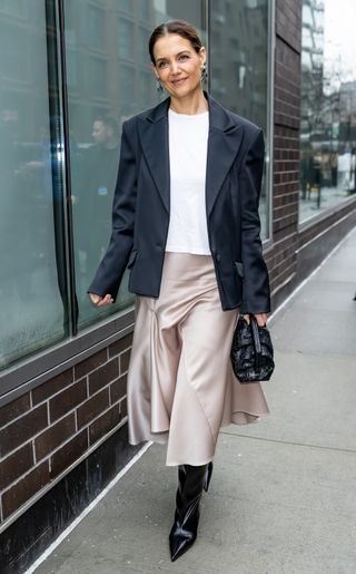 A photo of Katie Holmes pairing a flared taupe silk skirt with a navy blue blazer and slouchy black boots.