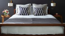 Dark grey bedroom with wooden double bed, cushions, white bedding, circular bedside tables with flowers, and two gold sconce lamps with white lampshades.