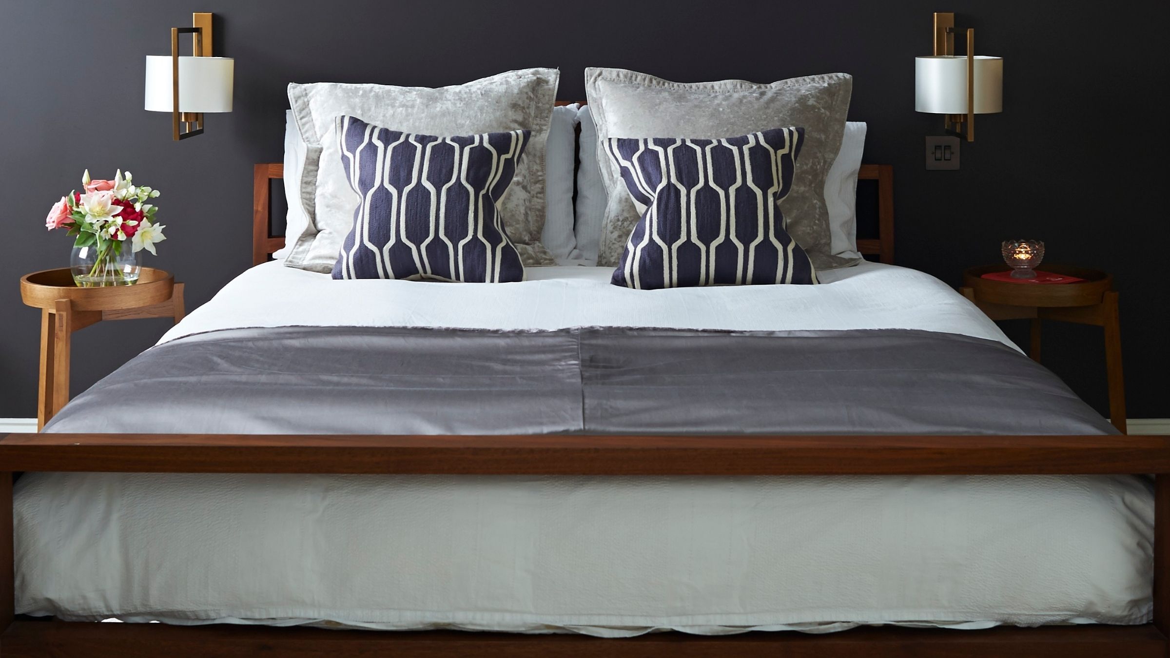 Dark grey bedroom with wooden double bed, cushions, white bedding, circular bedside tables with flowers, and two gold sconce lamps with white lampshades.