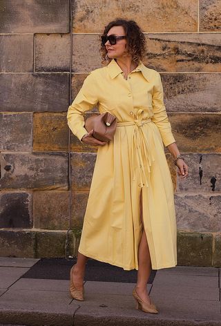 A woman wearing a yellow shirt dress with a tan clutch bag, tan heels, and brown sunglasses.