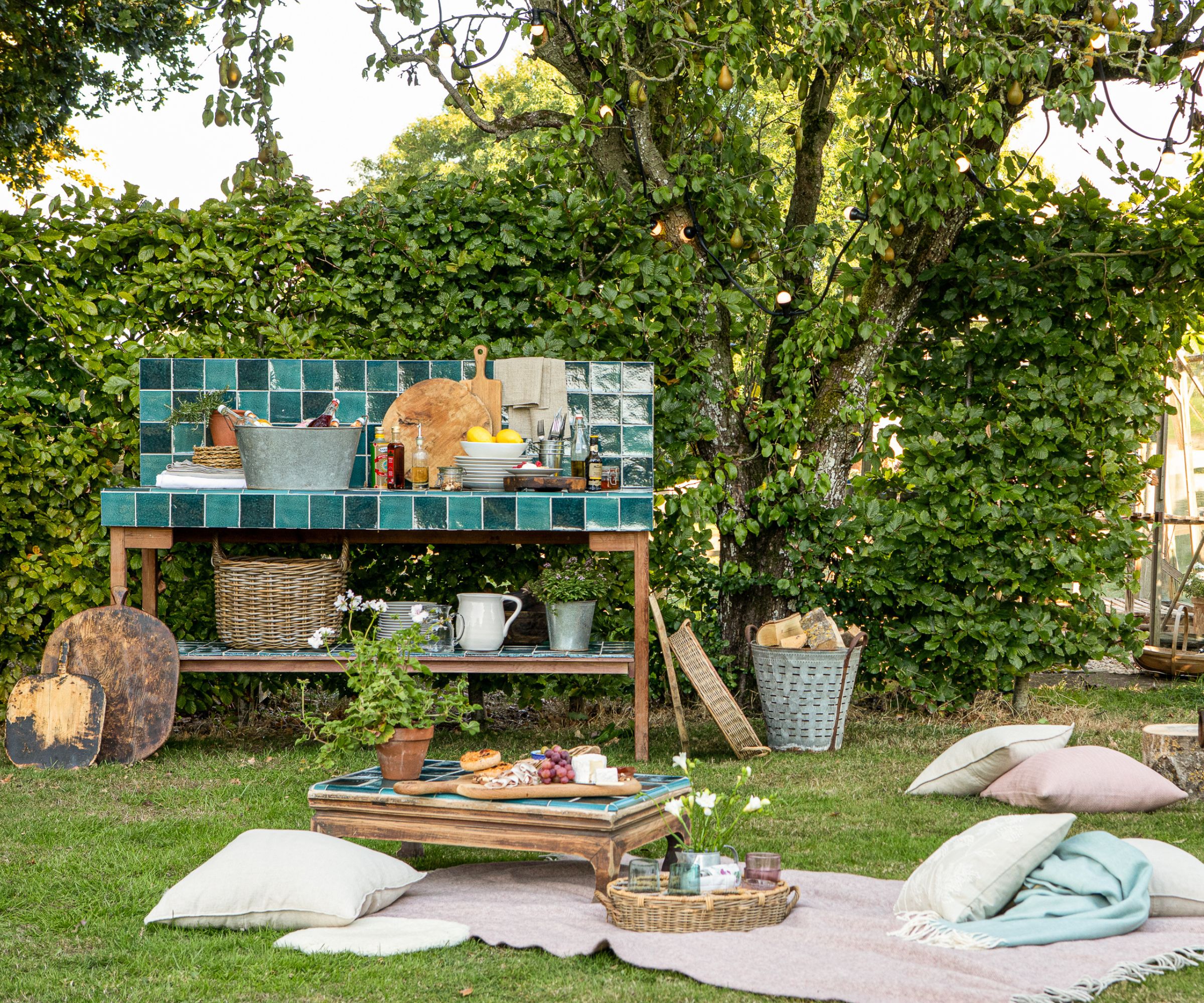 A garden party scene with a teal zellige-tiled outdoor prep station and matching low coffee table, floor cushions on a picnic blanket, wicker baskets and fairy lights.