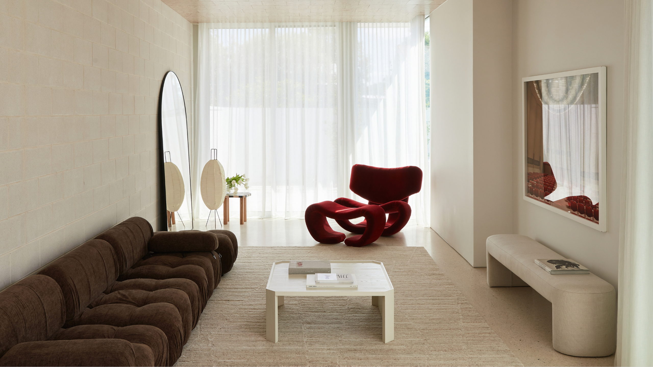 A white living room with a brown couch, a white coffee table, a bench, a framed print, an arched mirror, an area rug, a lantern, and a red accent chair