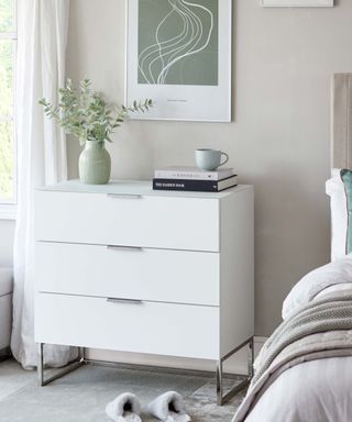 A white bedroom with a rectangular white set of drawers with a vase of green stems and books on top of it, a white and green rectangular wall art print above it, gray fluffy slippers below it and a gray bed next to it