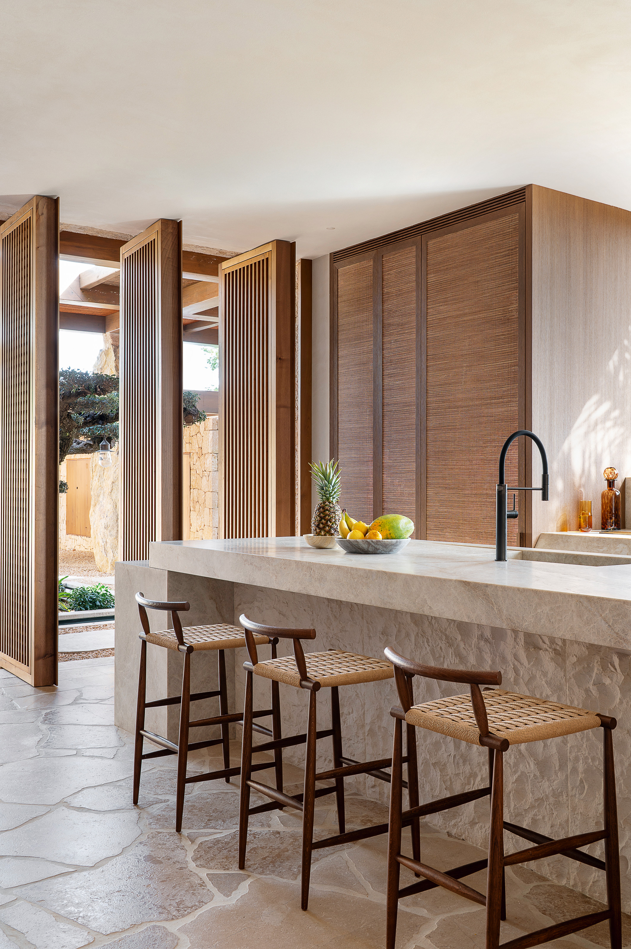 Kitchen with carved stone island with inset sink, wood and woven bar stools, wood cupboard with cane fronts