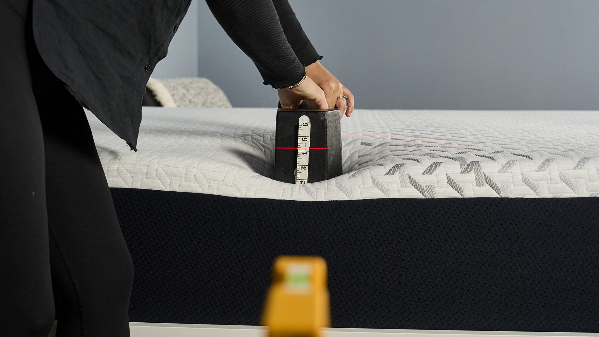 In this image we are preparing to perform an edge support test on the Nectar Premier Memory Foam Mattress. In the front of shot, out of focus, is a laser pen and level, directed at the surface of the Nectar Premier Mattress. A 25kg weight is being placed on the edge of the mattress by our tester Lauren Jeffries. There's a tape measure on the front of the weight and the laser is hitting the measure at roughly 4 inches, indicating how far the weight has sank into the mattress