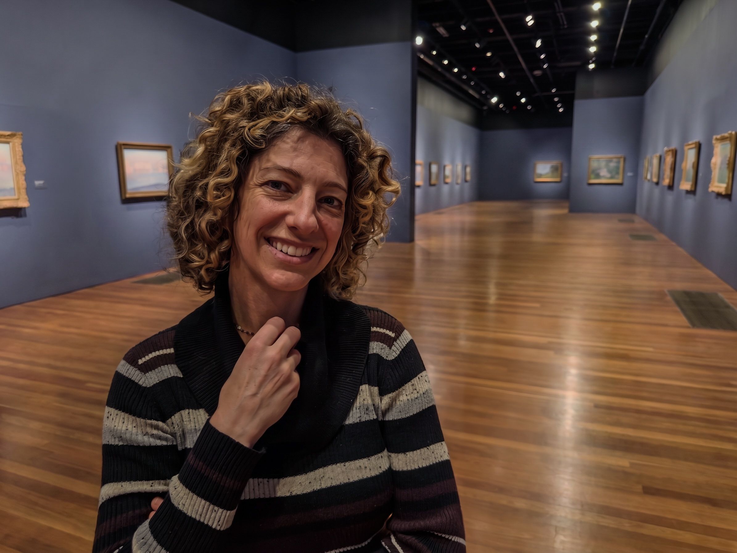 Smiling woman with curly hair poses inside an art gallery with framed paintings and polished wooden floors.