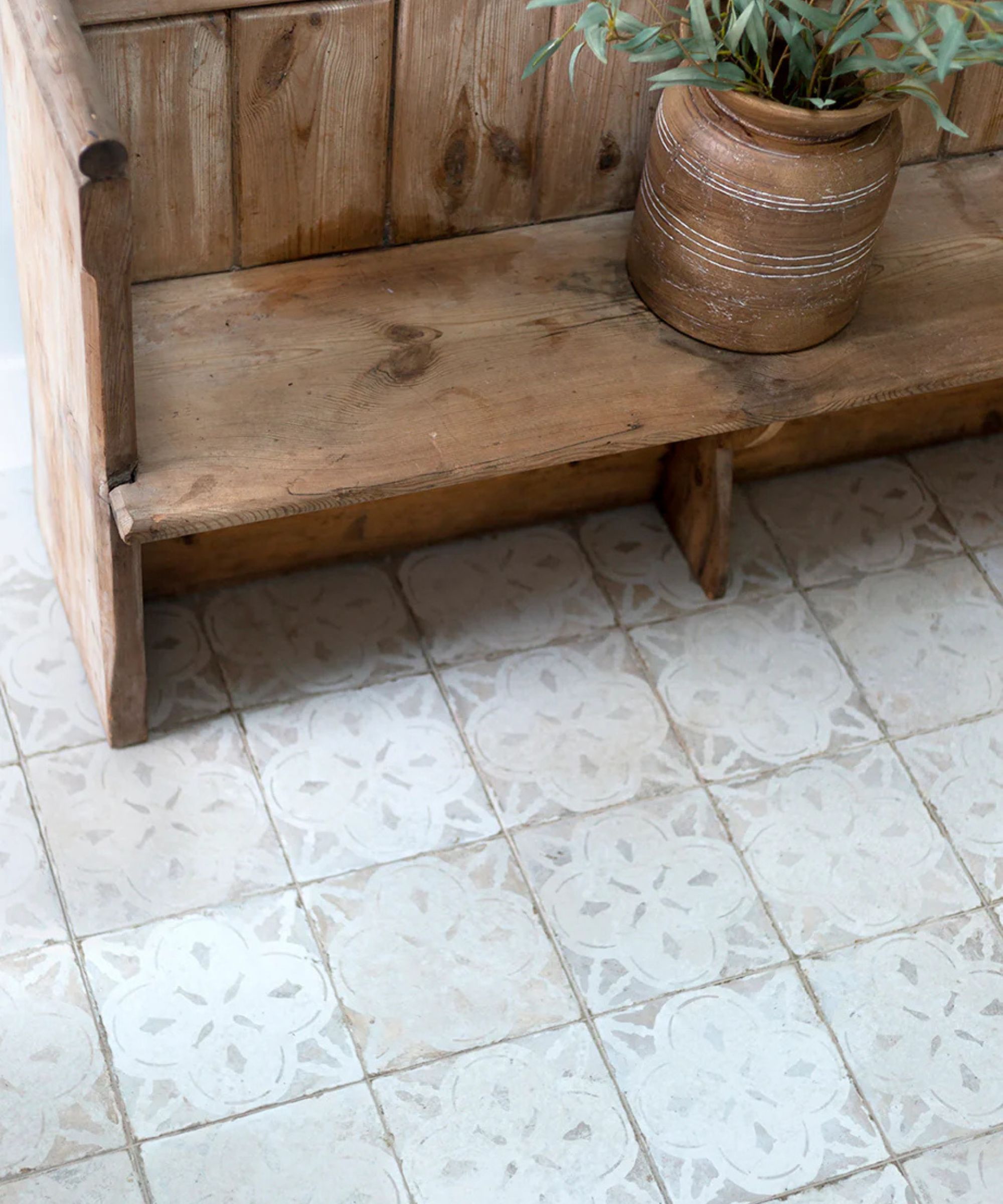 Rustic wooden bench with a weathered finish and a terracotta pot holding greenery. The floor features vintage-style, patterned square tiles in soft beige and white tones.