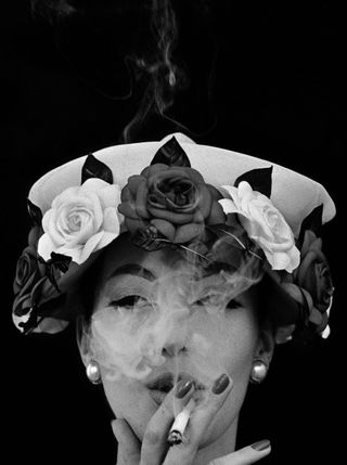 black and white image of a woman smoking a cigarette whilst wearing a hat adorned with 3 dark and 2 light coloured roses
