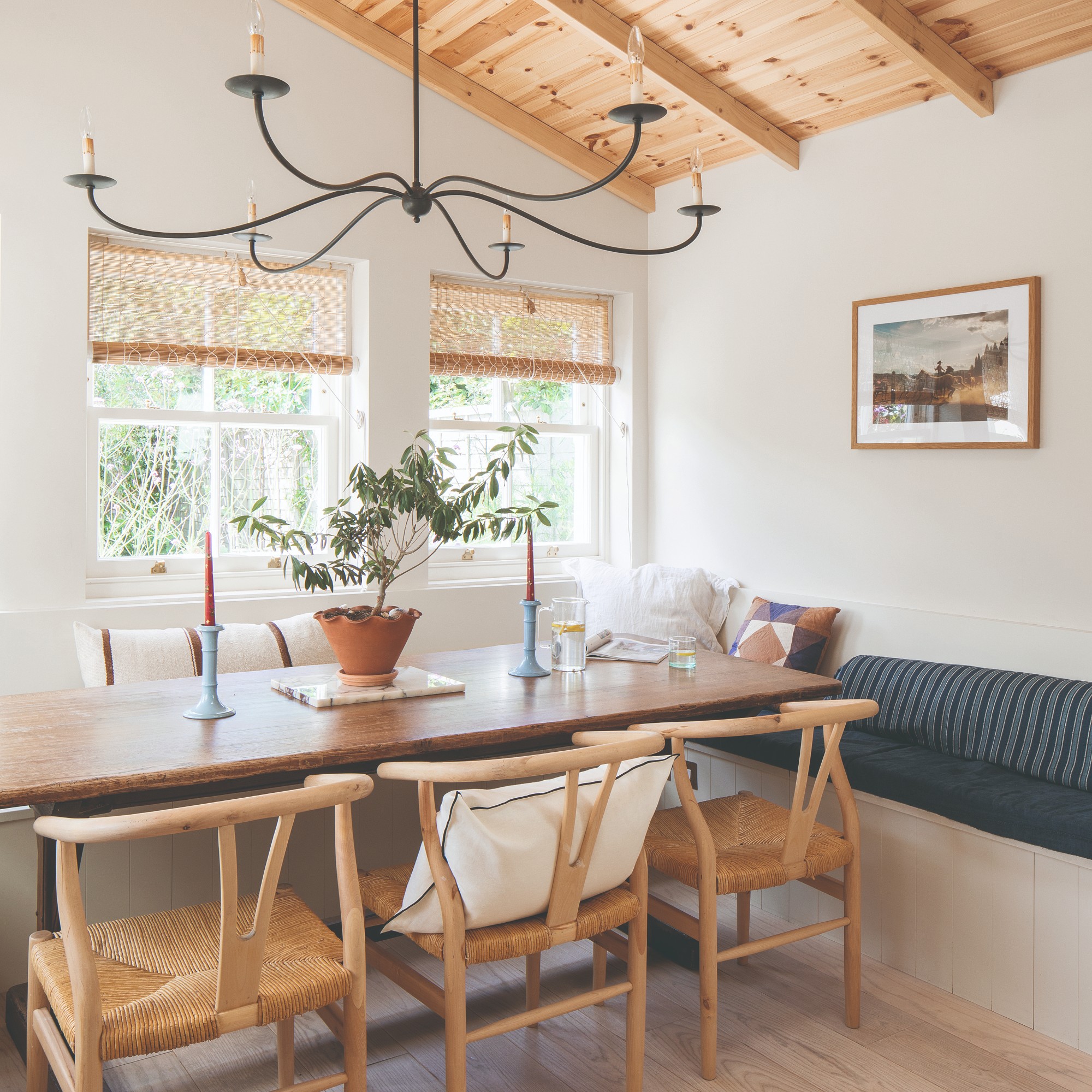 A dining room with wishbone chairs and a table decorated with a potted plant and two tapered red candles in light blue candle holders