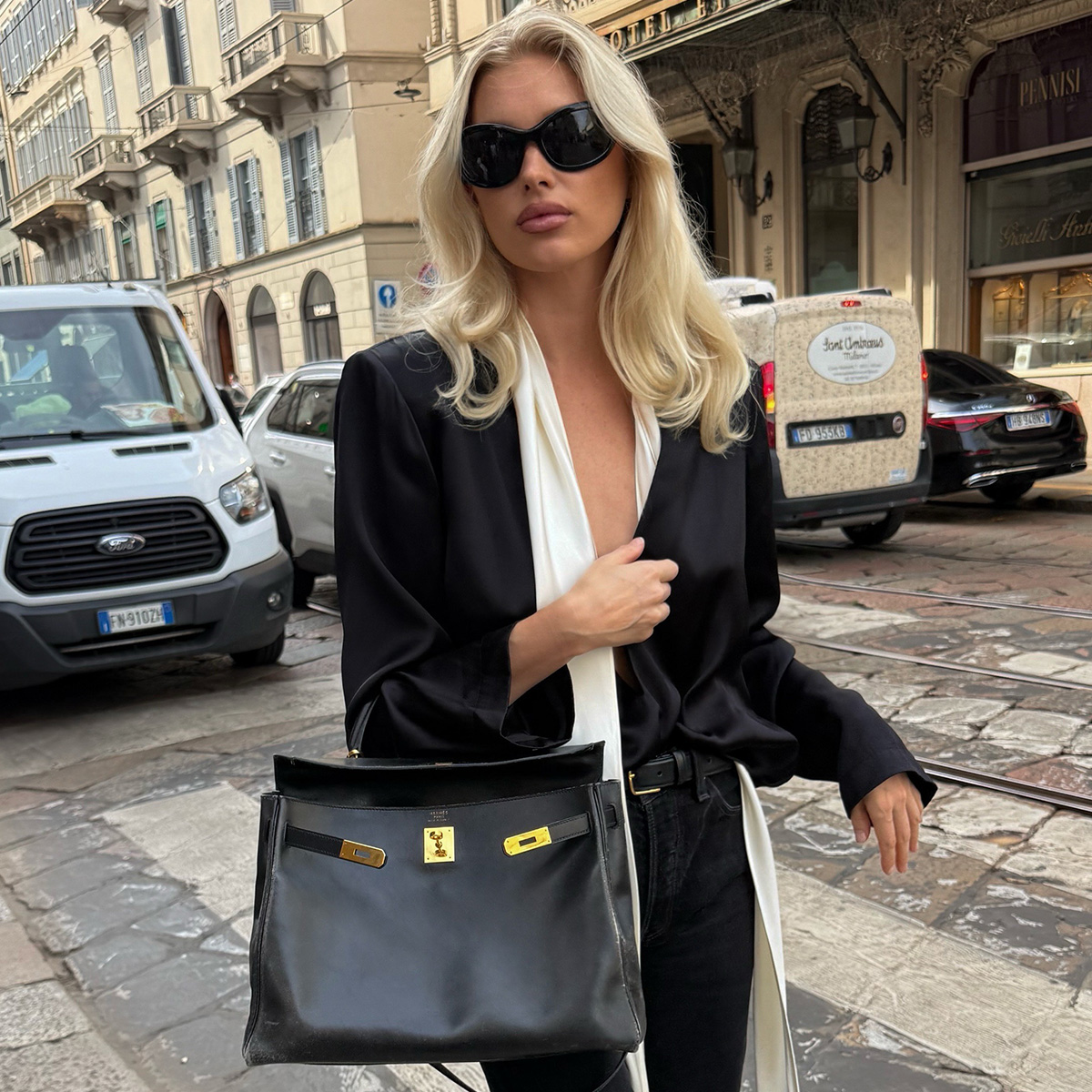 elsa wears oversize sunglasses, a black top, black jeans, and a white silk scarf.