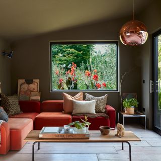 living room with large picture window with view of garden with orange flowers, orange sofa, dark olive green walls and copper-coloured light fitting