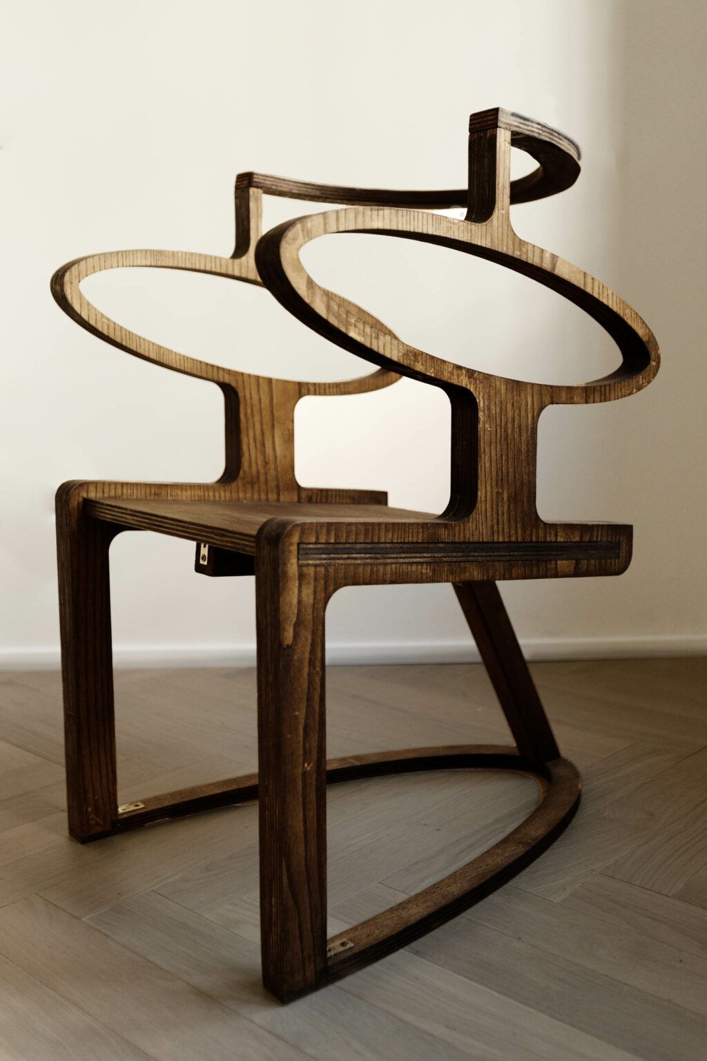 A futuristically designed wooden chair sits in a naturally lit room with wooden floors.