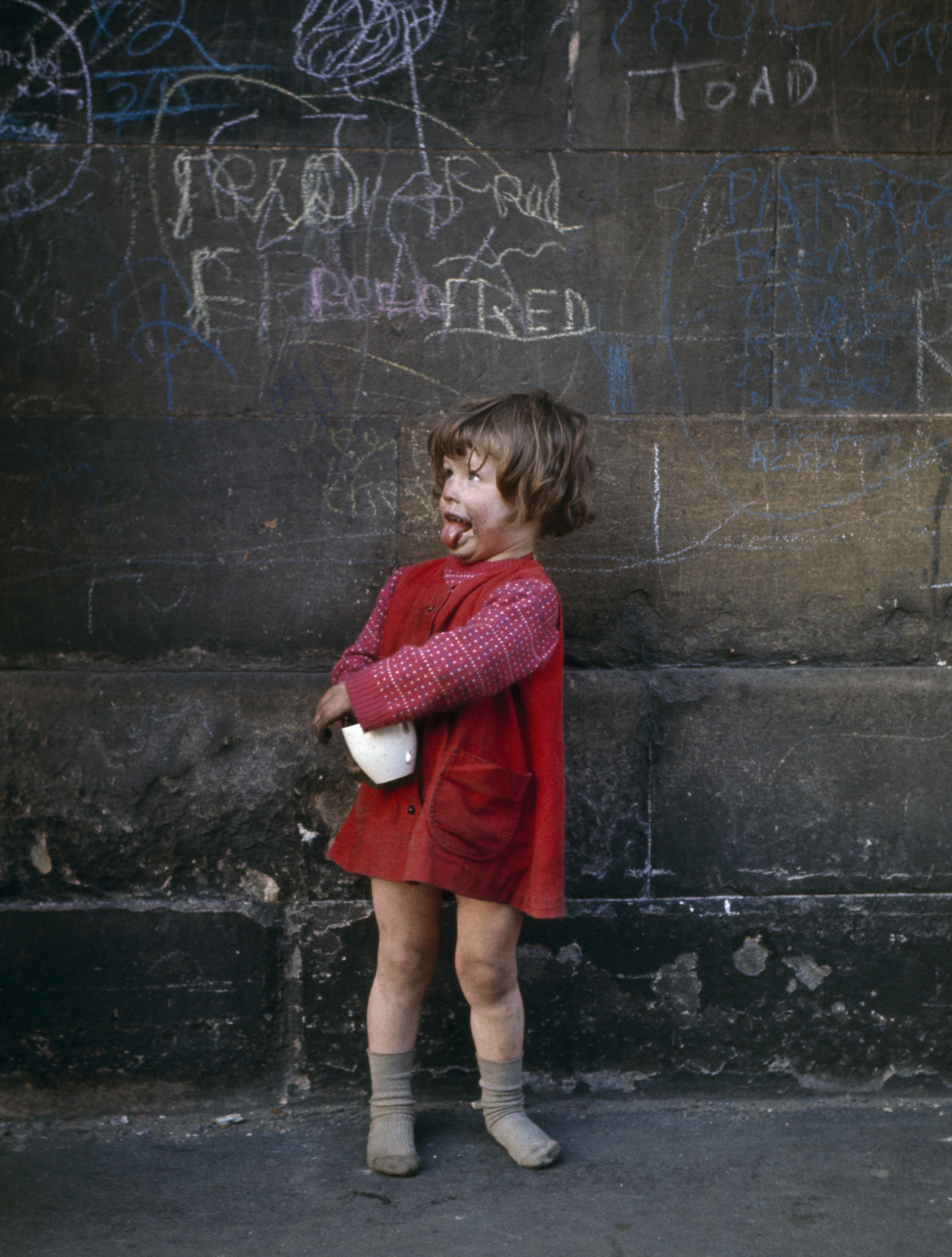 A young girl in a red dress and sweater, holding a white object, stands in front of a dark stone wall covered in white and blue chalk graffiti, sticking out her tongue.