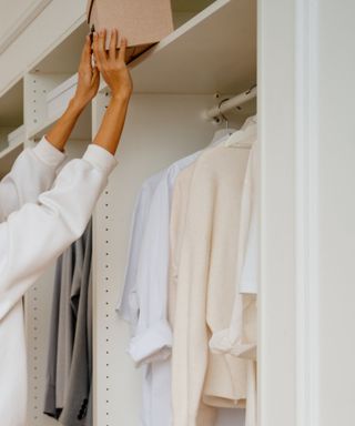 A pair of hands pushing a brown box into a white closet, with white shirts hanging inside of it