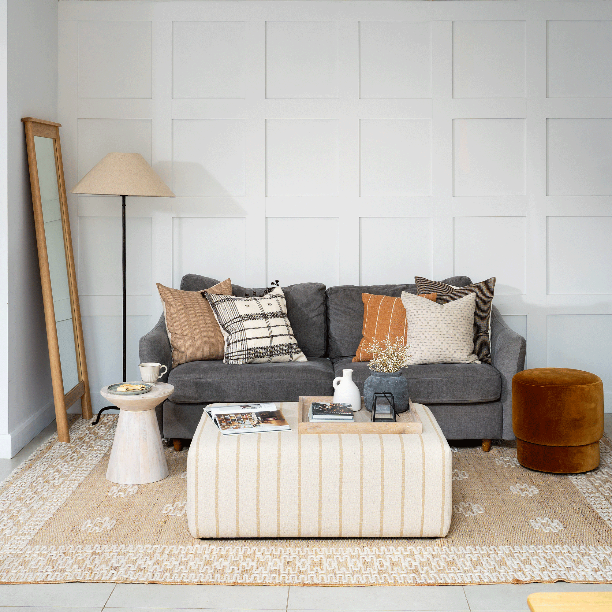 a living space with a dark grey sofa and striped ottoman a tall mirror and side table and white wall panelling